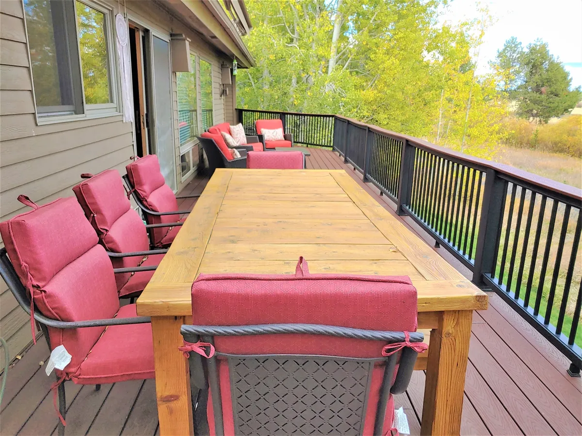 This exterior image features a wooden dining table surrounded by red cushioned chairs on a patio, bordered by a railing that offers views of the surrounding natural landscape. The setup invites guests to dine, relax, and enjoy outdoor gatherings in a tranquil environment.