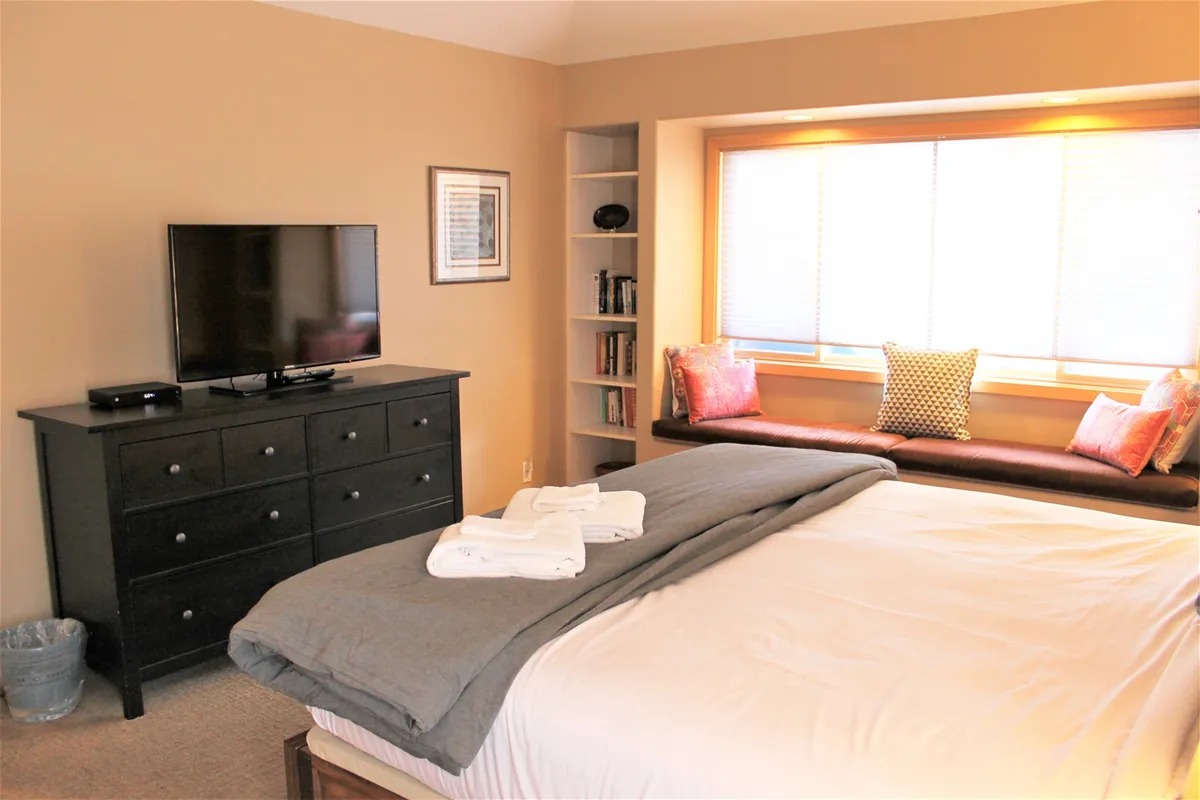 This image features a comfortable bed with soft bedding, a television mounted nearby, and a window seat that admits plenty of natural light. The setup invites moments of relaxation, reading, or winding down with a show, creating an atmosphere of tranquility and restful retreat.