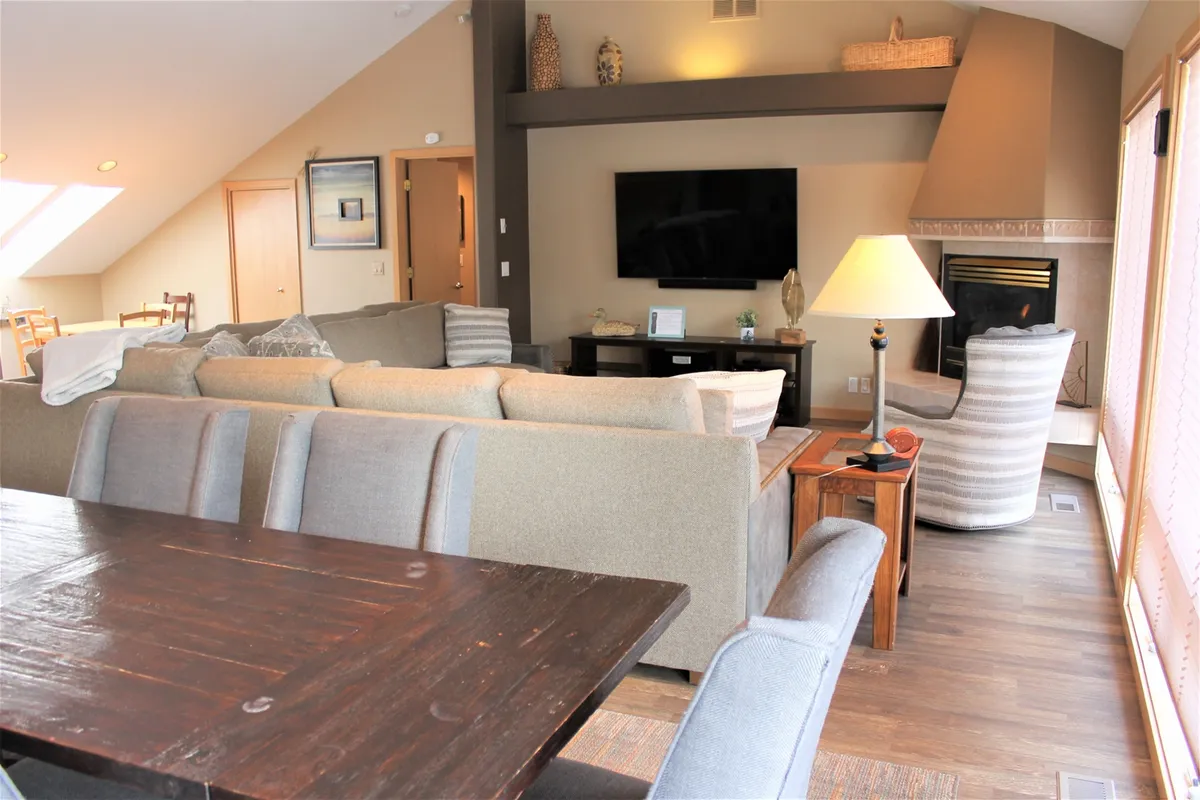 A warm and welcoming living room featuring a large sectional sofa, elegant fireplace, dining table, and a television. The room is designed for group interactions, cozy relaxation by the fire, and shared meals, all in a single inviting space.