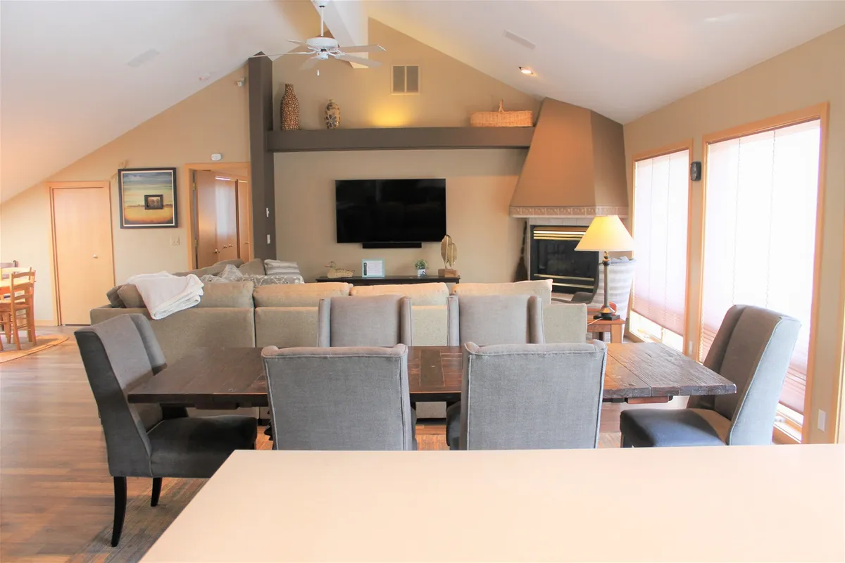 The image shows a spacious, sunlit living area featuring a comfortable sofa, a modern dining table, a cozy fireplace, and a large television. The room exudes warmth and relaxation, perfect for family gatherings, movie nights, and unwinding after a day of adventure. Inviting touches and updated décor create a welcoming atmosphere ideal for groups and families.