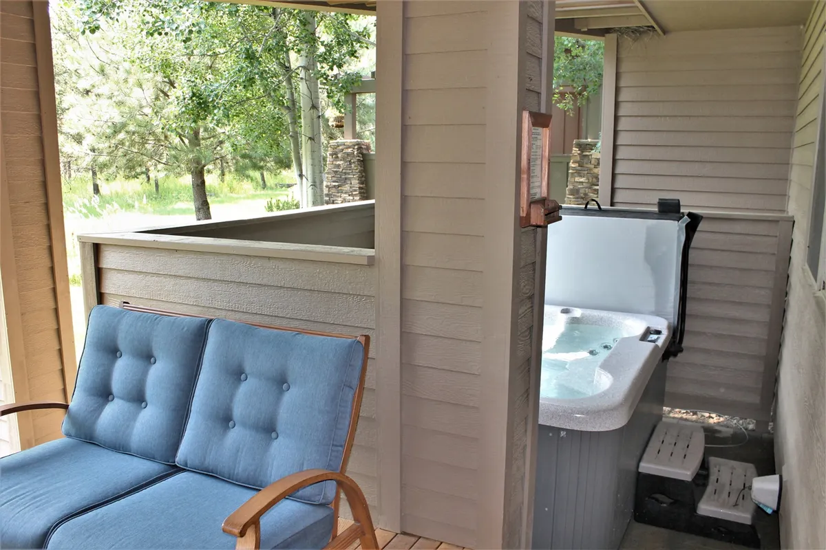 A serene outdoor space featuring a blue cushioned sofa and a hot tub set against vibrant greenery. The setup invites both relaxation and socializing, with the natural backdrop highlighting the connection between indoor comforts and the great outdoors.
