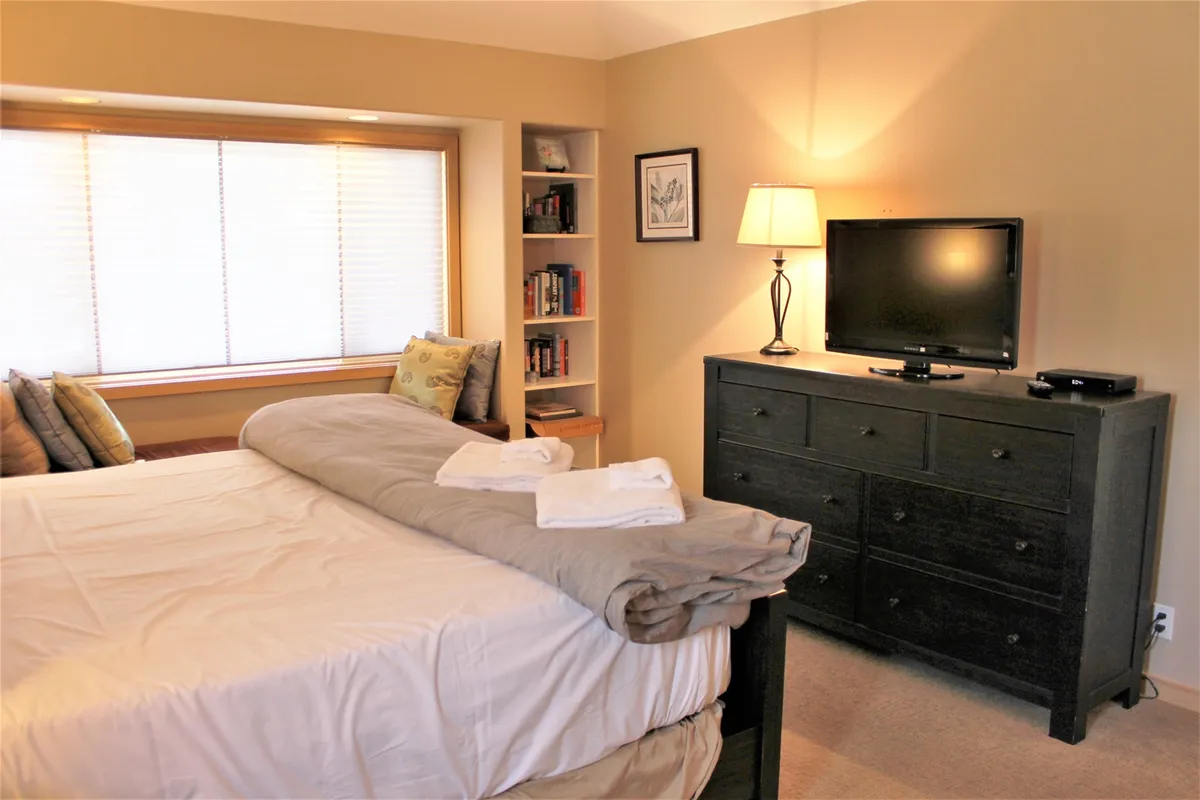 A serene bedroom with a bed facing a window with a cozy seating nook, a bookshelf, and a dresser with a television. The room is bathed in natural light, providing a versatile space for relaxation, reading, or watching TV.